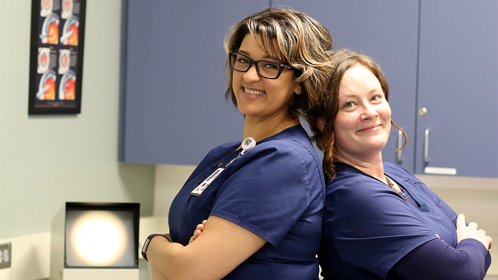 Two students in scrubs standing back to back and smiling at the camera
