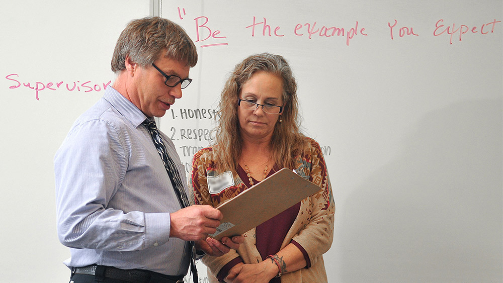 Instructor and a student looking at a clipboard in front of a whiteboard that says 'be the example'